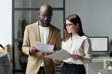Young serious businessman and brunette woman looking through financial documents while discussing papers