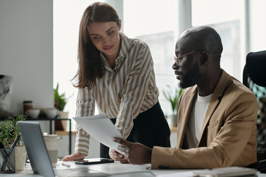 Young businesswoman bending over workplace and looking at financial paper document held by male colleague - Powered by Adobe