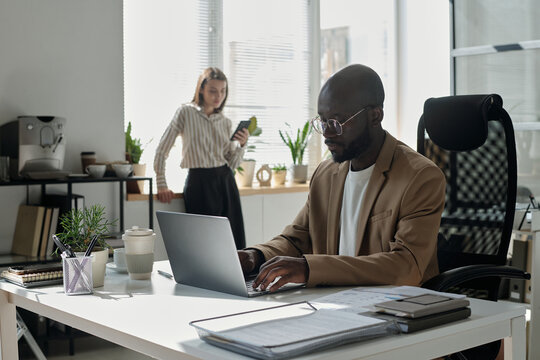 Serious young male analyst touching keys of laptop keypad while working with online data against colleague
