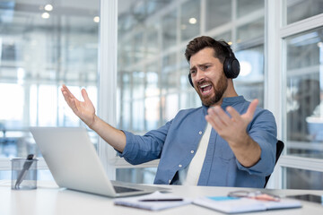 Dissatisfied man sits in headphones inside the office at his workplace, employee looks at laptop...