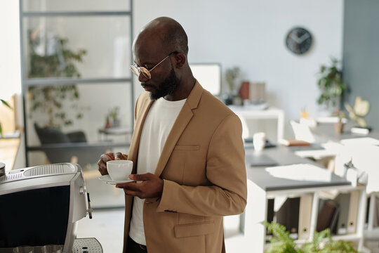 Serious young male ceo in formalwear holding cup of aromatic drink while standing in front of coffee machine
