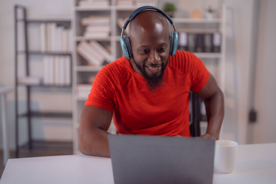 Young african american businessman wearing a red t-shirt is smiling while having a video call on his laptop using headphones in his home office
