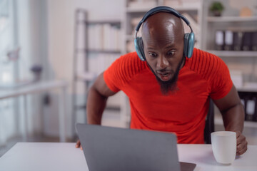 Surprised African American man wearing headphones, working from home, sipping coffee, and staring...