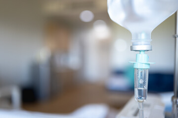 Close-up of an IV drip in a hospital room, with blurred medical equipment and furniture in the background.