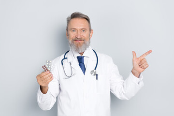 Senior male doctor in white coat holding blister packs of pills with a smile, pointing to the side...
