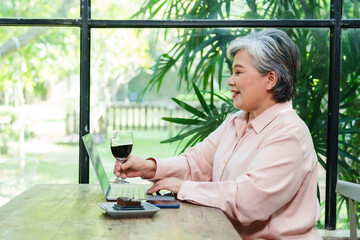 Fototapeta premium Elderly Asian woman relaxing after retirement enjoying coffee while working on laptop at cafe. Senior embracing digital lifestyle, balancing productivity and leisure in a cozy environment.