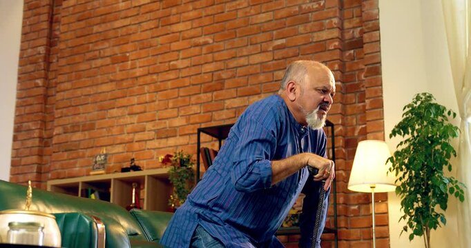 Indian elderly retired man with long beard wearing kurta, struggling to stand up with a walking stick, sitting on a sofa in a modern living room with helpless expressions and visible effort