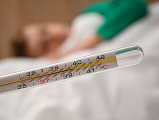 Mercury thermometer showing high body temperature of 39.6 degrees celsius with sick child laying in bed in the background