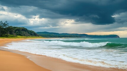 Obraz premium Moody Seascape with Waves Crashing on Sandy Beach Under Dramatic Cloudy Sky and Lush Green Hills in Background
