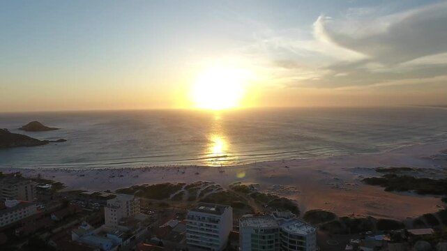  p&ocirc;r do sol na praia de Arraial do Cabo, transformando o c&eacute;u em uma paleta de cores vibrantes, com tons de laranja, rosa e dourado refletindo nas &aacute;guas cristalinas. O drone sobrevoa suavemente a paisa