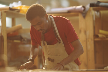 An African American craftsman wearing an apron, diligently examining his woodworking project in a...