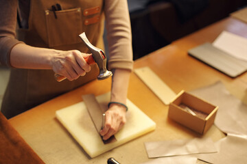 Person meticulously crafting leather goods on workspace with various tools. One hand holding...