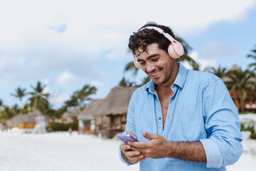 hispanic young man using headphones and listening music on phone in vacations or holidays at the beach in Mexico Latin America, Caribbean and tropical destination	