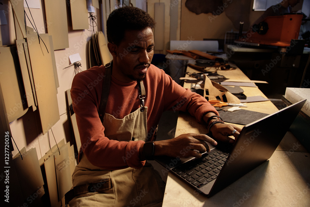 Poster man engaged in leather crafting project in a dimly lit workshop, utilizing laptop for design referen