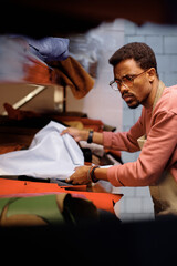 Man wearing glasses and apron focusing on crafting leather goods in workshop, with various tools...