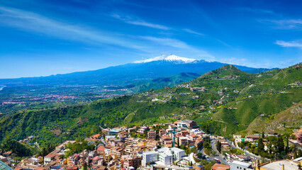 Panoramic view of Taormina, historic town in Sicily, Italy, surrounded by lush green hills. In distance, Mount Etna, highest active volcano in Europe with its snow-capped peak