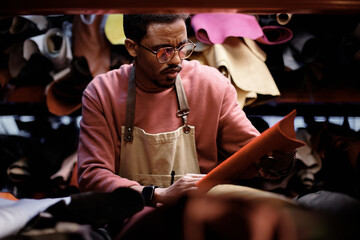 Craftsman wearing apron inspecting piece of leather in a workshop filled with various leather materials. Focused, meticulous work environment with shadows creating depth