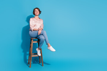 Young woman in stylish satin blouse sitting on high stool against vibrant blue background