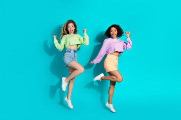 Two cheerful women in colorful outfits jumping with joy against a vivid turquoise background