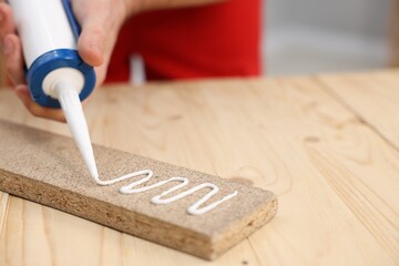 Man with caulking gun glueing wooden plank at table, closeup