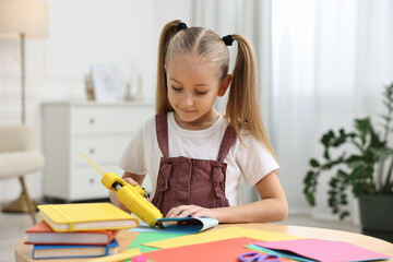 Little girl with hot glue gun and color paper making craft at table indoors