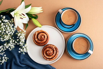 Cinnamon rolls on a white plate with two cups of coffee in blue ceramic cups, white lilies, and gypsophila flowers arranged on a beige background with a dark blue fabric. Top view, flat lay