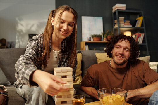 Pretty girl removing block from the center of tower built on table while playing tabletop game with boyfriend
