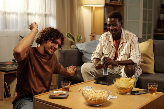Happy guy with playing card in hand looking at poker on table during tabletop game with his friend
