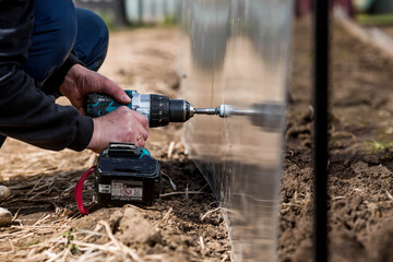 A man holds a battery-powered screwdriver and assembles a new greenhouse made of polyethylene,...