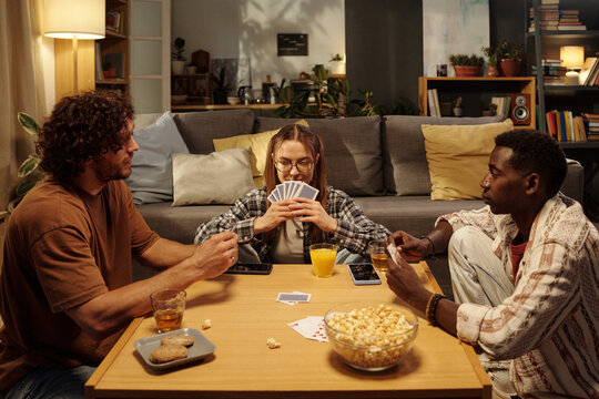 Two intercultural guys and girl with playing cards sitting by table with snacks and drinks during tabletop game