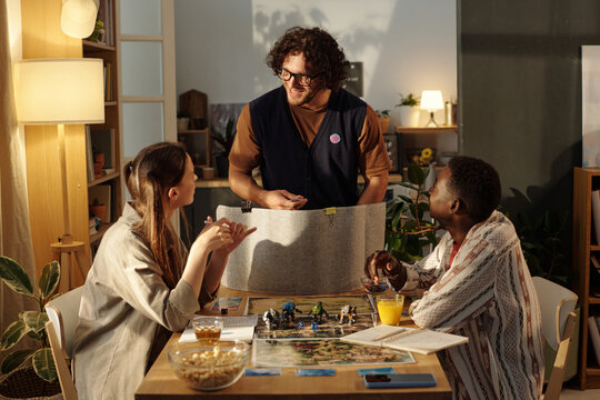 Happy young man looking at one of participants of tabletop game and explaining rules before playing
