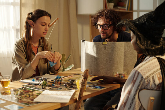 Young female goblin sitting by table and looking at tabletop game picture during play with two male friends