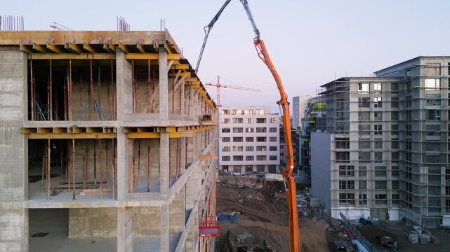 Aerial view of an active construction site featuring a high-rise building under development. A concrete boom pump pours cement onto the rooftop, with cranes, scaffolding, and structural framework