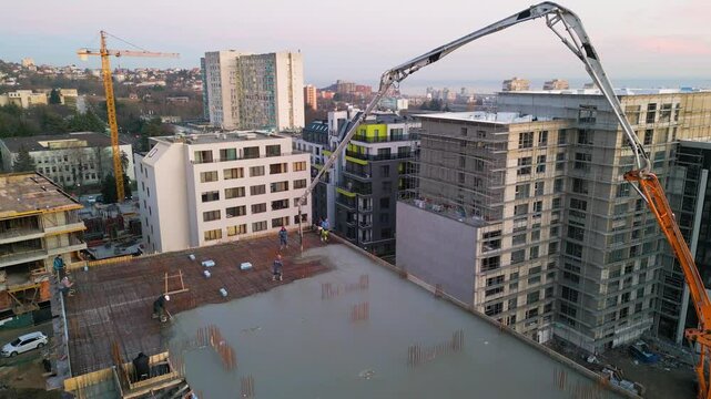 Aerial view of a construction site with a high-rise building under development. A concrete pump pours cement on the rooftop, while workers and scaffolding indicate progress. Surrounded by modern