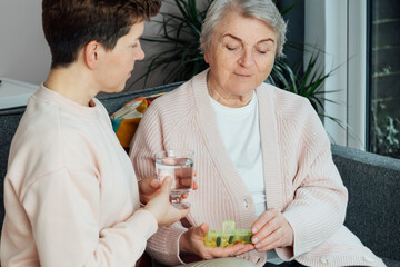 Middle aged neutral gender woman taking care of old mother or grandma, giving pills or supplements and glass of water helping taking medicine on time. Elderly healthcare and support. Age caregiving.