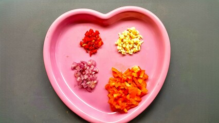 A pink heart-shaped plate containing four types of chopped ingredients: red onion, red chili, garlic, and tomato. It is interesting because it shows the preparation of the ingredients.