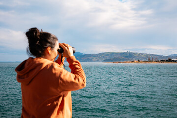 Woman in an Orange Hoodie Using Binoculars to Observe Coastal Landscape in Santoña, Cantabria, Spain During Springtime
