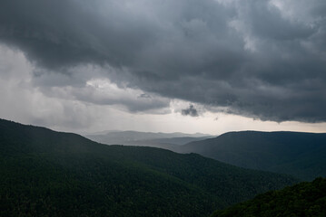 This image captures a dramatic mountain landscape under a heavy, stormy sky. Dark, textured clouds dominate the frame, casting a moody and somewhat ominous atmosphere over the scene. 