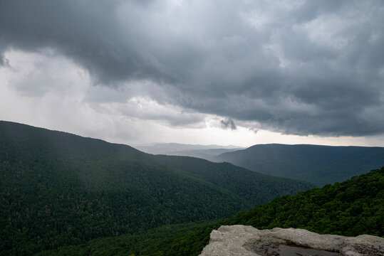 This image captures a dramatic mountain landscape under a heavy, stormy sky. Dark, textured clouds dominate the frame, casting a moody and somewhat ominous atmosphere over the scene. 