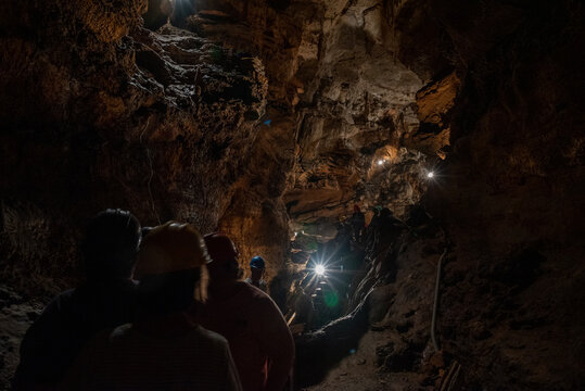 A group of people wearing helmets are exploring a dimly lit cave with rugged rock formations. The cave is illuminated by several bright lights