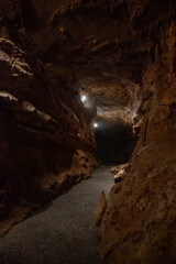 the interior of a dimly lit cave with a gravel path running through it. The cave walls and ceiling are rugged and textured, with small stalactites hanging from the ceiling. 