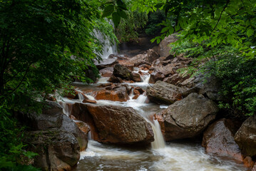A lush forest setting features a cascading stream flowing over large rocks. The greenery surrounding the water adds a serene atmosphere to the scene.
