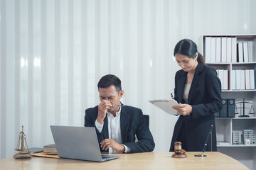 Fototapeta premium Legal Stress: A young lawyer looks overwhelmed and stressed at his desk while his assistant looks on, capturing the pressure and workload of a legal career. The image features a laptop, legal books.