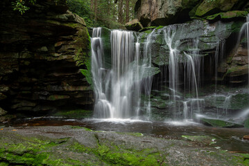 A serene waterfall cascades over moss-covered rocks in a lush forest setting. The gentle flow of water creates a tranquil atmosphere 