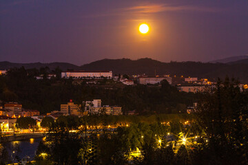A bright full moon illuminates the night sky over a cityscape with illuminated buildings, a winding river, and surrounding hills.