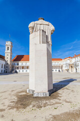 Obraz premium A large stone monument in the form of a draped figure stands in a spacious square with historic buildings and a clock tower under a bright blue sky.