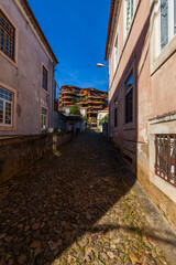 A narrow cobblestone street leads uphill towards a modern building complex with curved lines, contrasting old and new architecture under a bright blue sky.