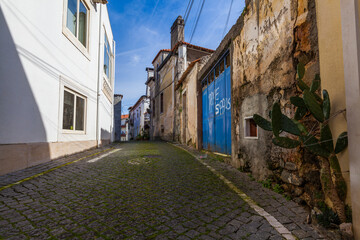A narrow cobblestone street lined with old buildings, featuring weathered walls, a bright blue gate with graffiti, and a touch of greenery, under a bright blue sky.