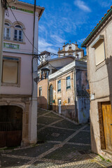 A narrow cobblestone street winds uphill between old buildings, offering a glimpse of a church tower against a bright blue sky with wispy clouds.