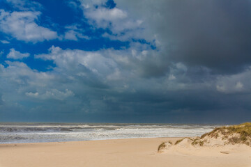 A dramatic sky with dark storm clouds contrasting against patches of blue hangs over a sandy beach and a turbulent ocean with visible waves.